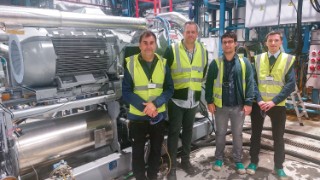 Vier Personen stehen nebeneinander in einer Werkshalle. Sie tragen Warnschutzwesen und Sicherheitsschuhe und blicken direkt ins Bild. Links hinter ihnen sieht man eine neue technische Anlage.