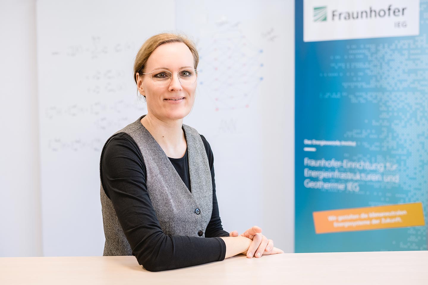 Tanja Kneiske ist Scientific Manager Integrierte Energieinfrastrukturen beim Fraunhofer IEG Eine Frau mit hellen Haaren und Brille sitzt an einem Schreibtisch. Sie trägt eine graue Weste und darunter ein schwarzes T-Shirt mit langen Armen. Im Hintergrund sieht man eine weiße Wand mit mathematischen Formeln und ein Banner vom Fraunhofer IEG